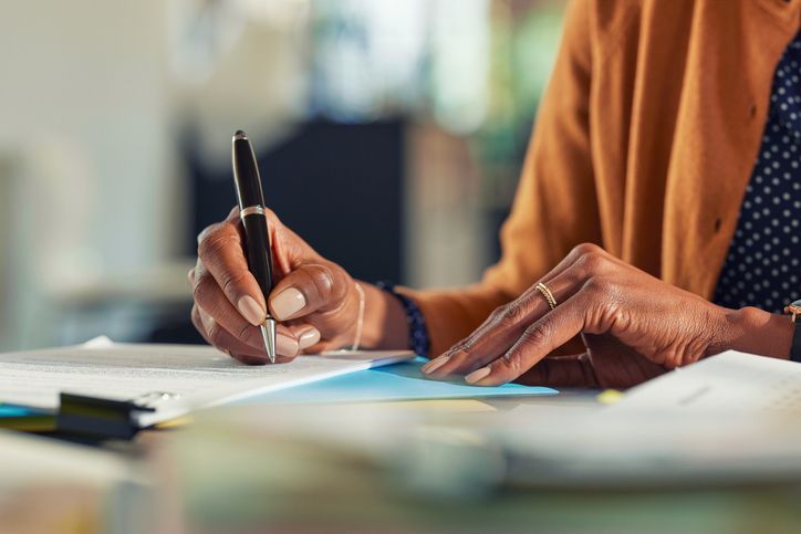 Person writing with a pen at a desk, wearing a tan sweater and a ring.