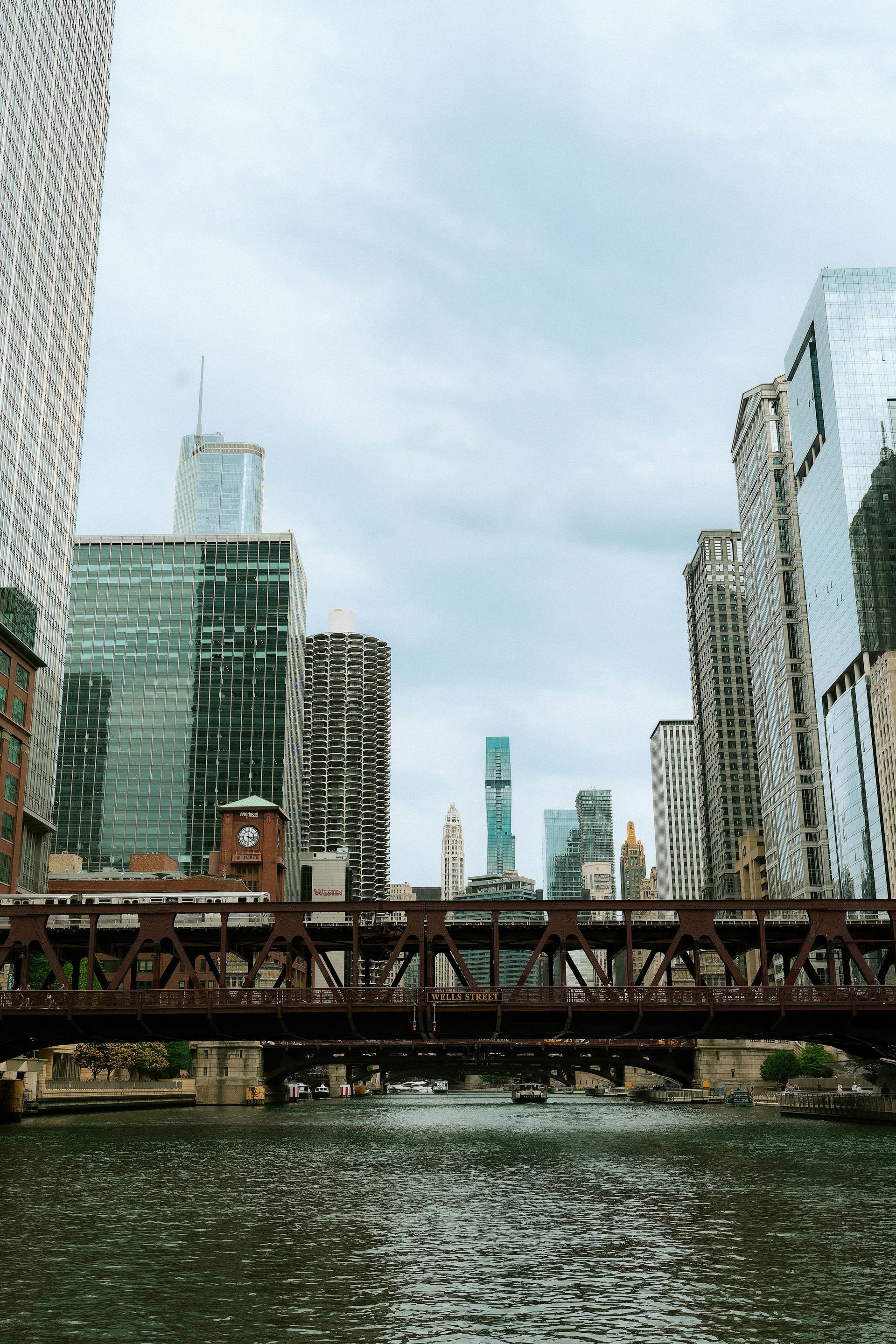 Chicago skyline framed by a dark metal bridge over a river, with cloudy sky overhead.