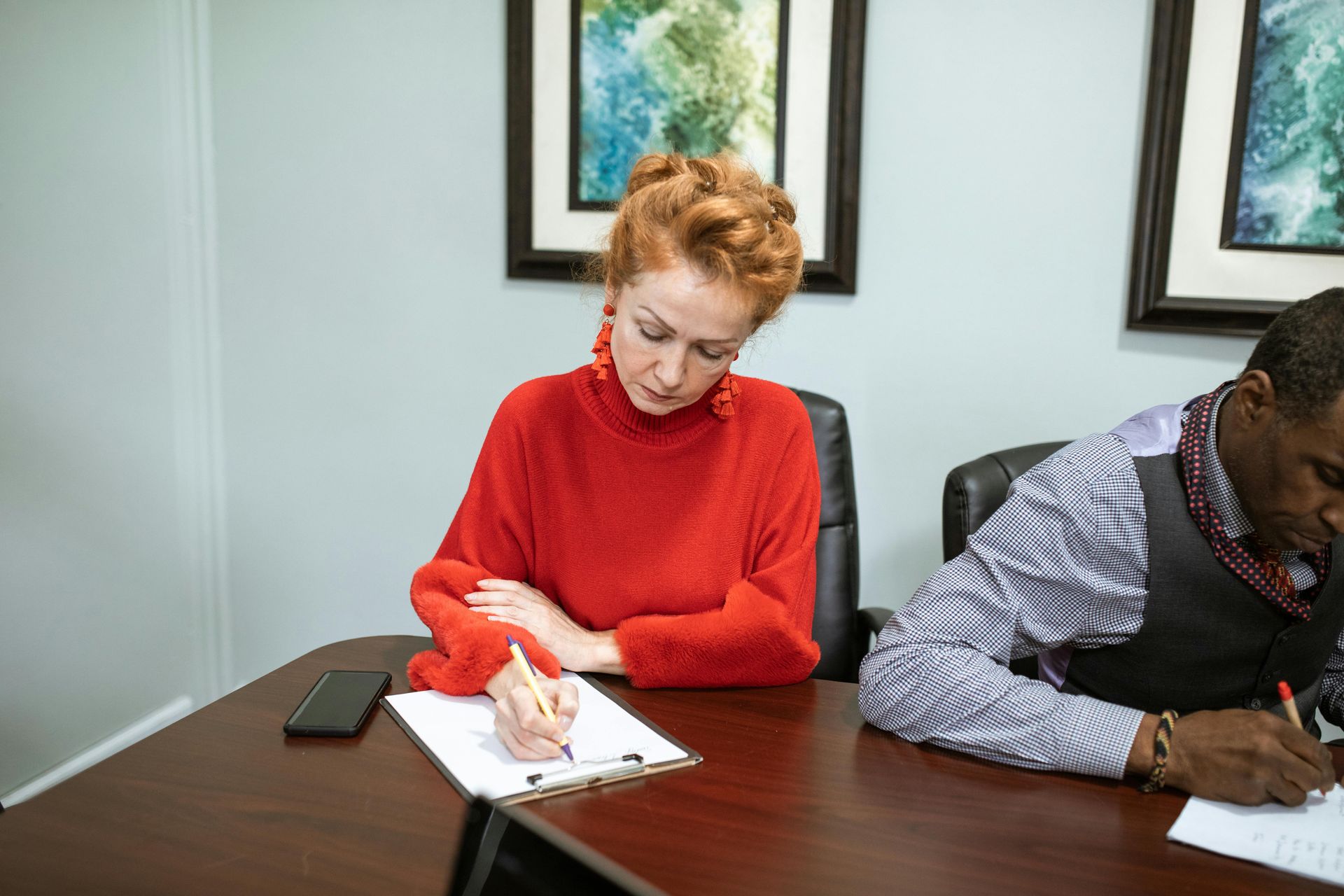 Woman in red sweater and man in striped shirt writing at a desk, looking down.