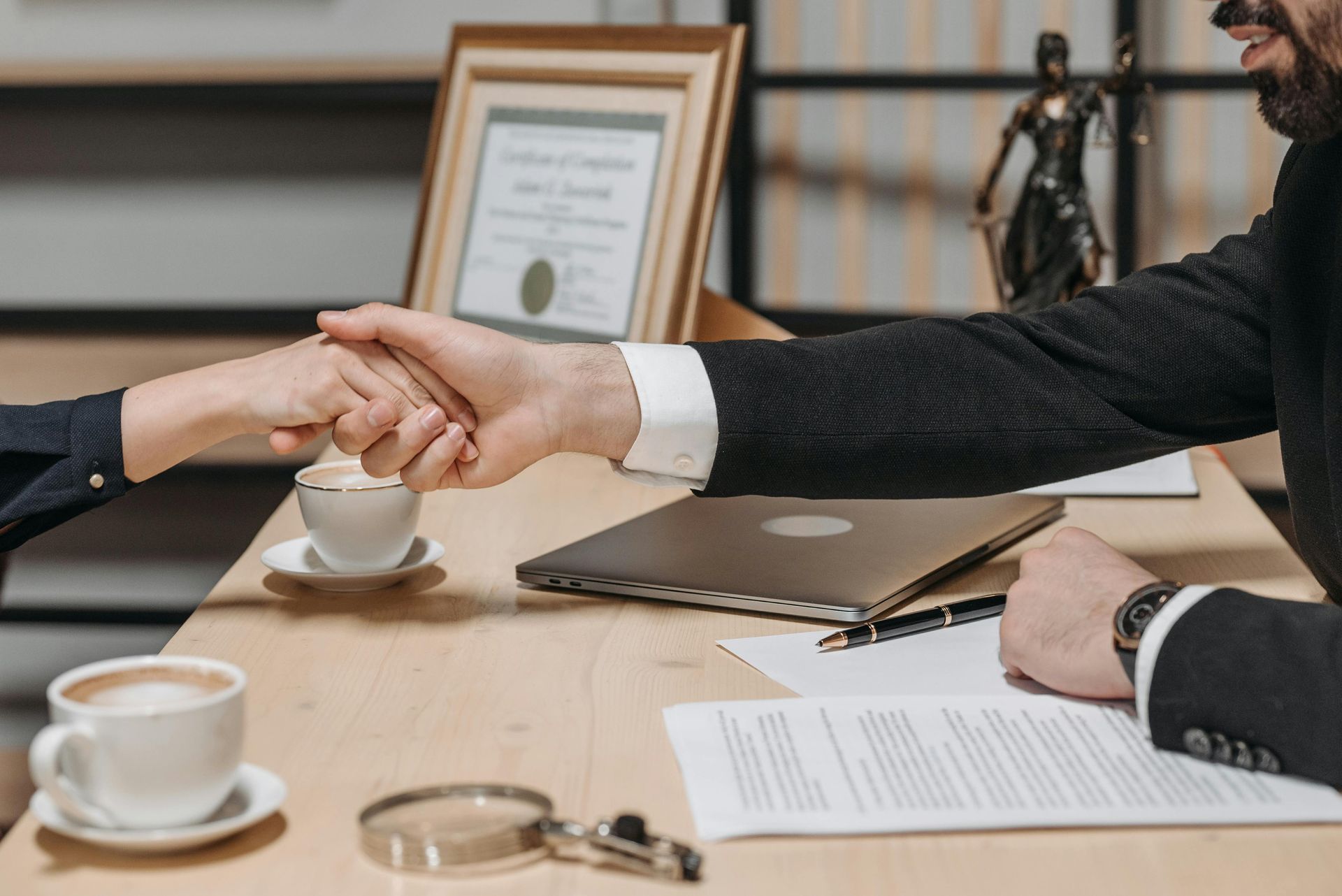 Two people shaking hands over a desk with documents, coffee cups, and a laptop.