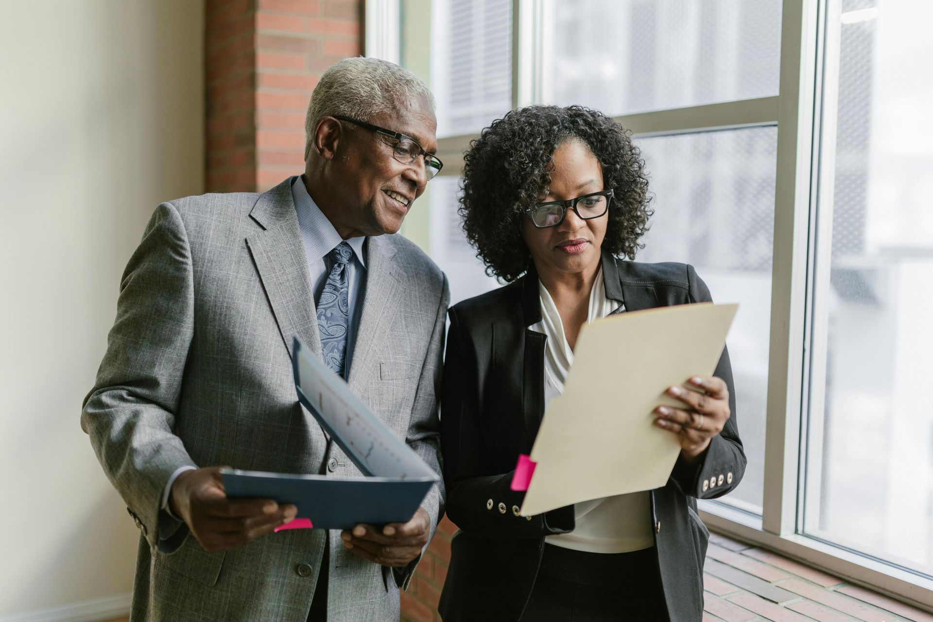 Two people reviewing documents near a window. Man in gray suit smiles. Woman wears a black blazer, glasses.