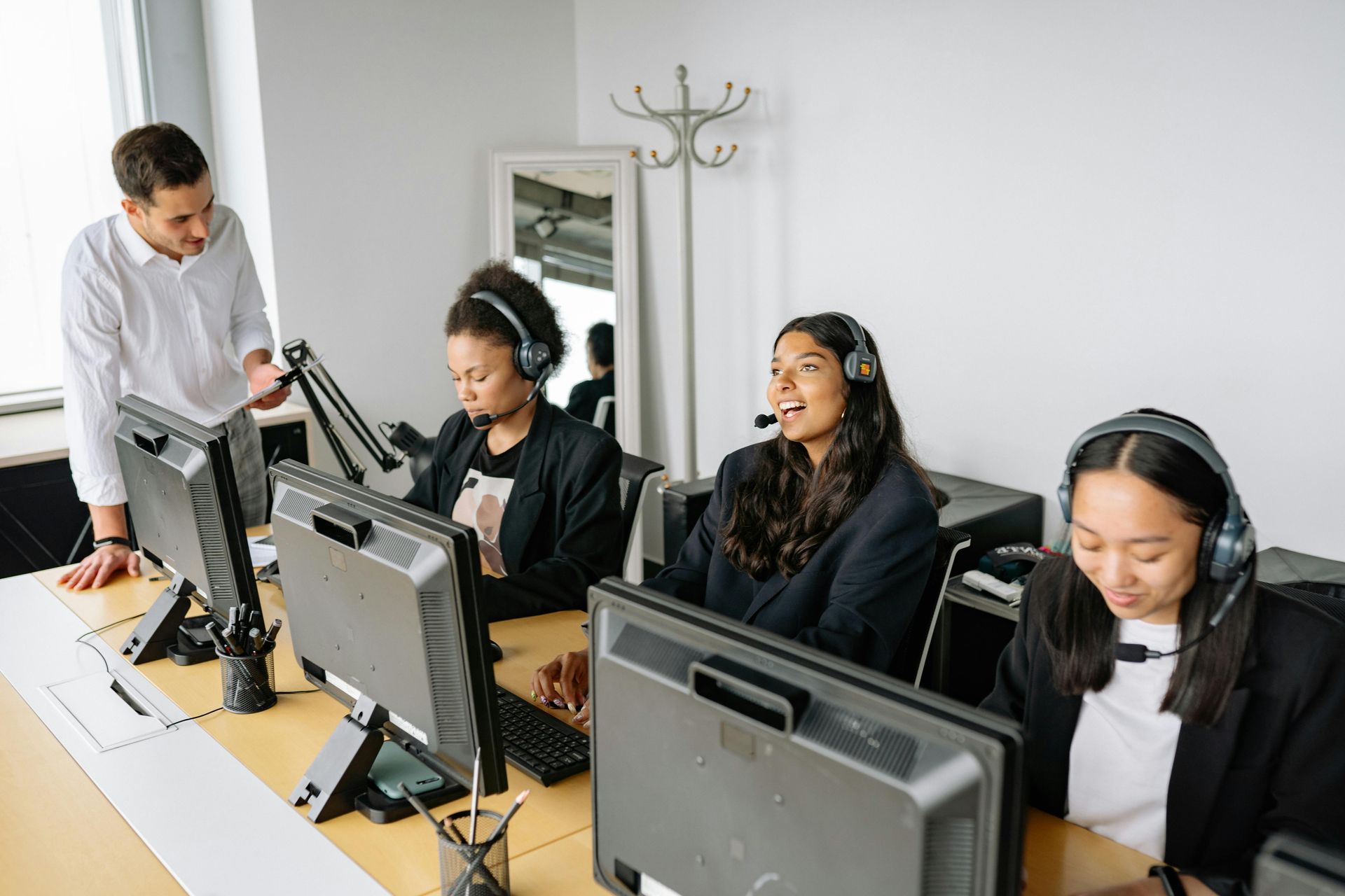 Office setting: manager assisting three employees wearing headsets at computers.
