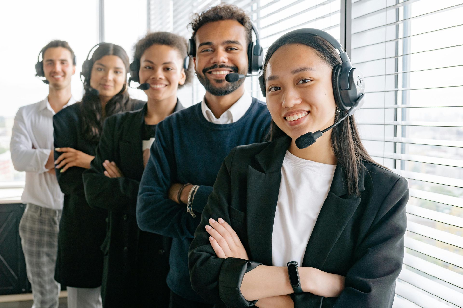 Five people in headsets smile, arms crossed, in a bright office near a window.