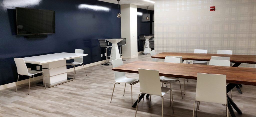 A room with tables and chairs. Dark blue wall with TV and white tables. Long wooden tables with white chairs.