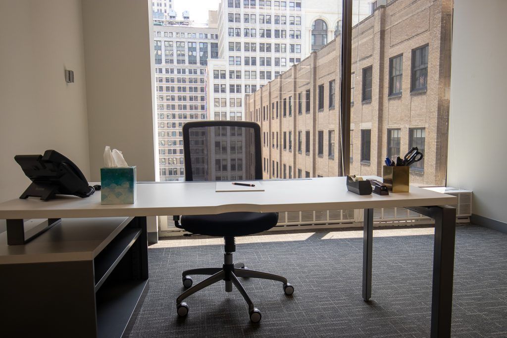 Office with desk, chair, tissue box, phone, and pens, facing a city view through a large window.