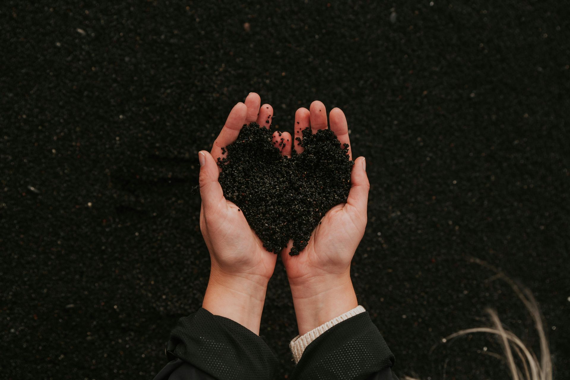 Hands holding black sand in a heart shape against a dark, sandy background.