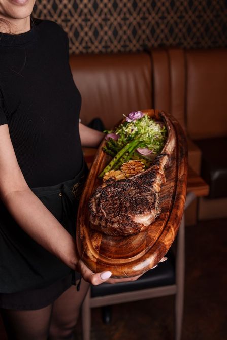 A woman is holding a wooden cutting board with a large piece of meat on it.