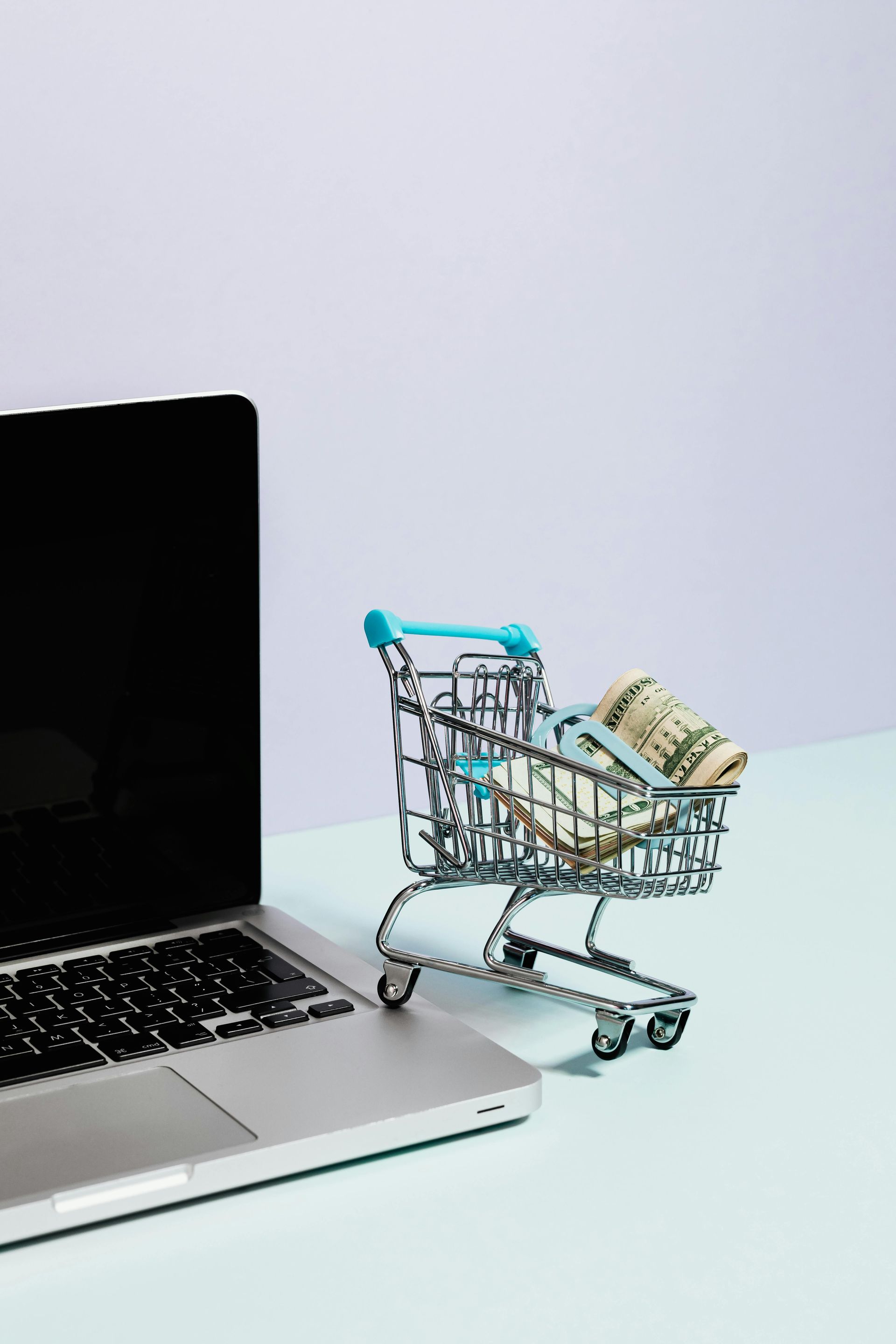 A shopping cart filled with money is next to a laptop computer.