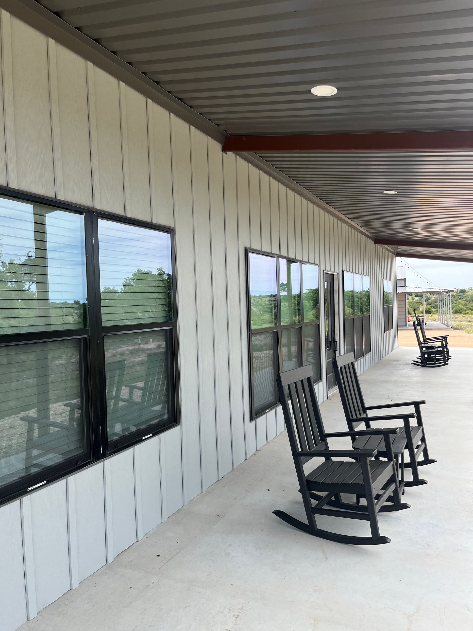Exterior view of a building with white siding, black-framed windows, and rocking chairs on a covered porch.