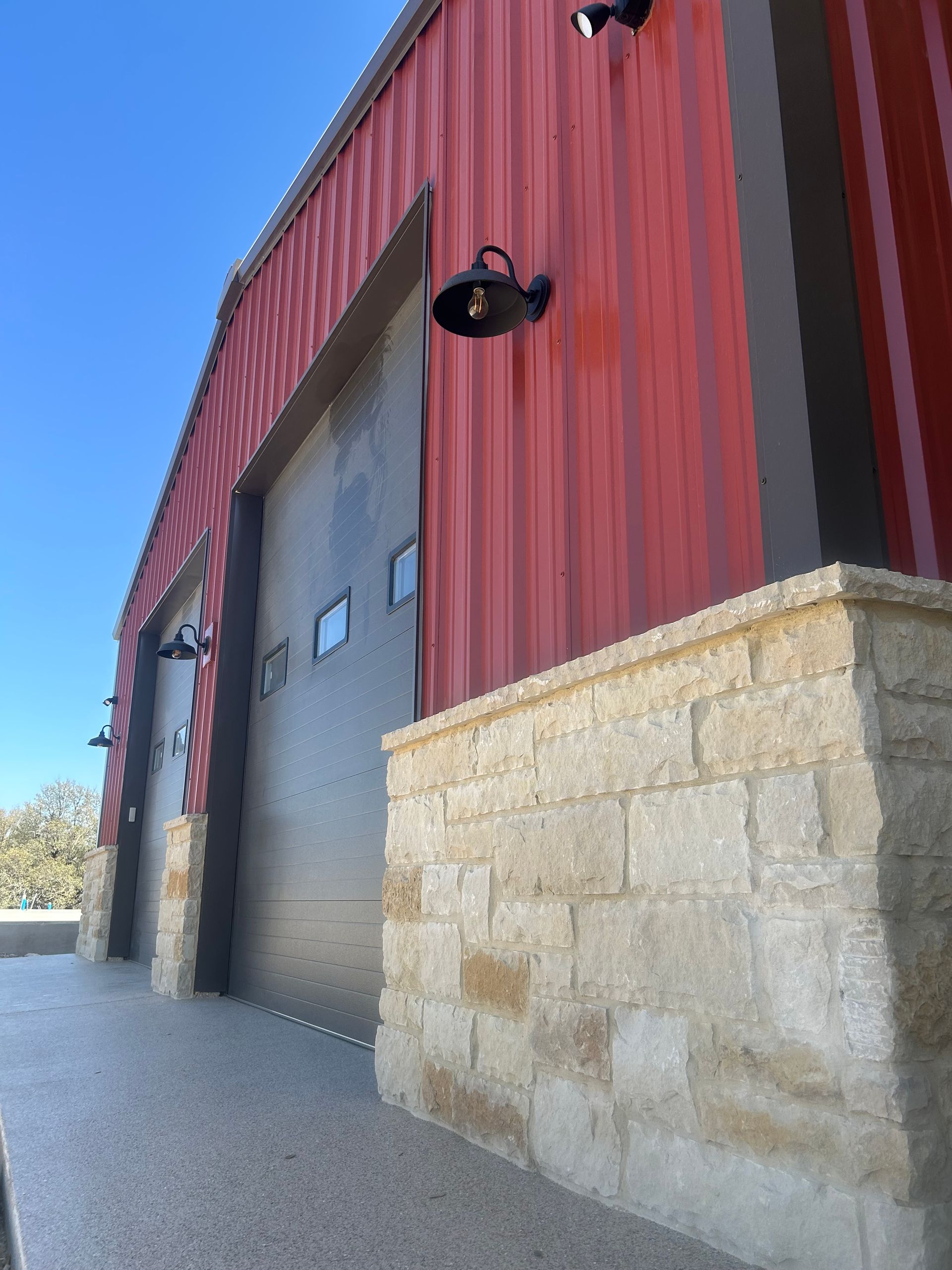 Red metal building with garage doors, stone accents, and exterior lighting against a blue sky.