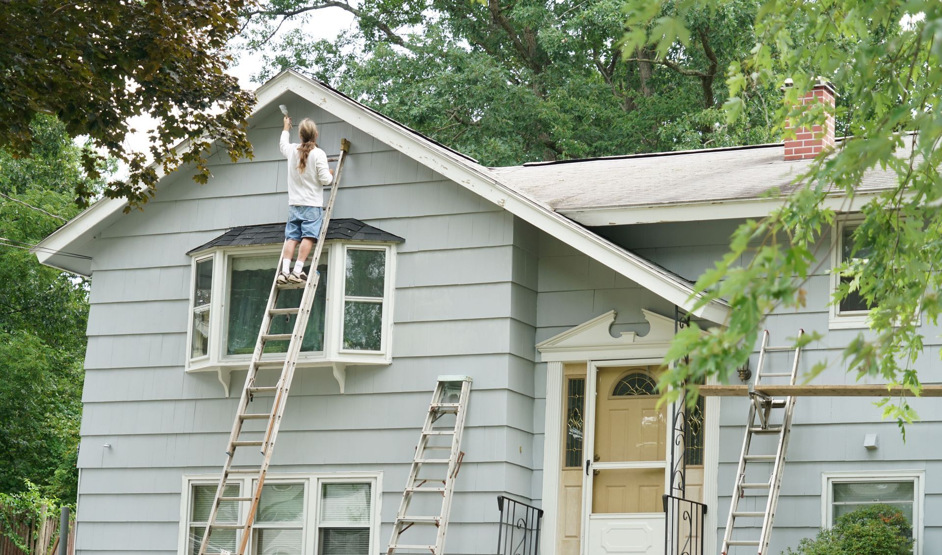 Person standing on a ladder repainting the exterior trim of a two-story house.