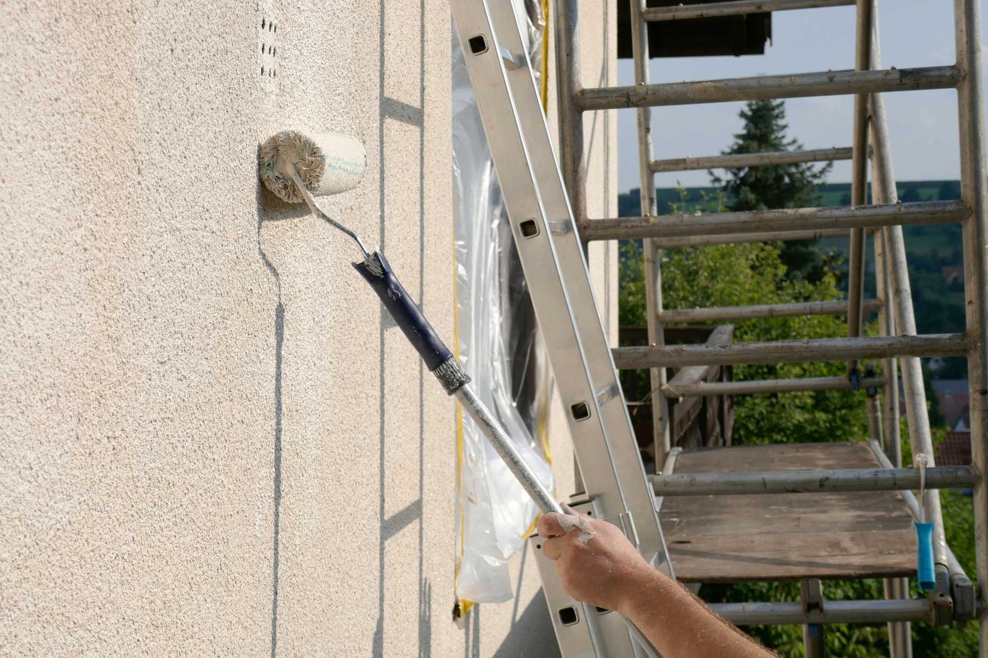 Contractor applying primer to exterior stucco wall with paint roller beside ladder and scaffolding.