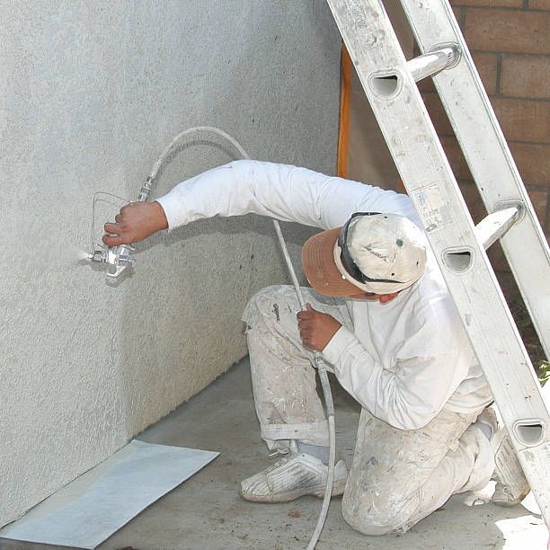 Male worker wearing a white uniform spray paints the exterior of a stucco wall with white paint.