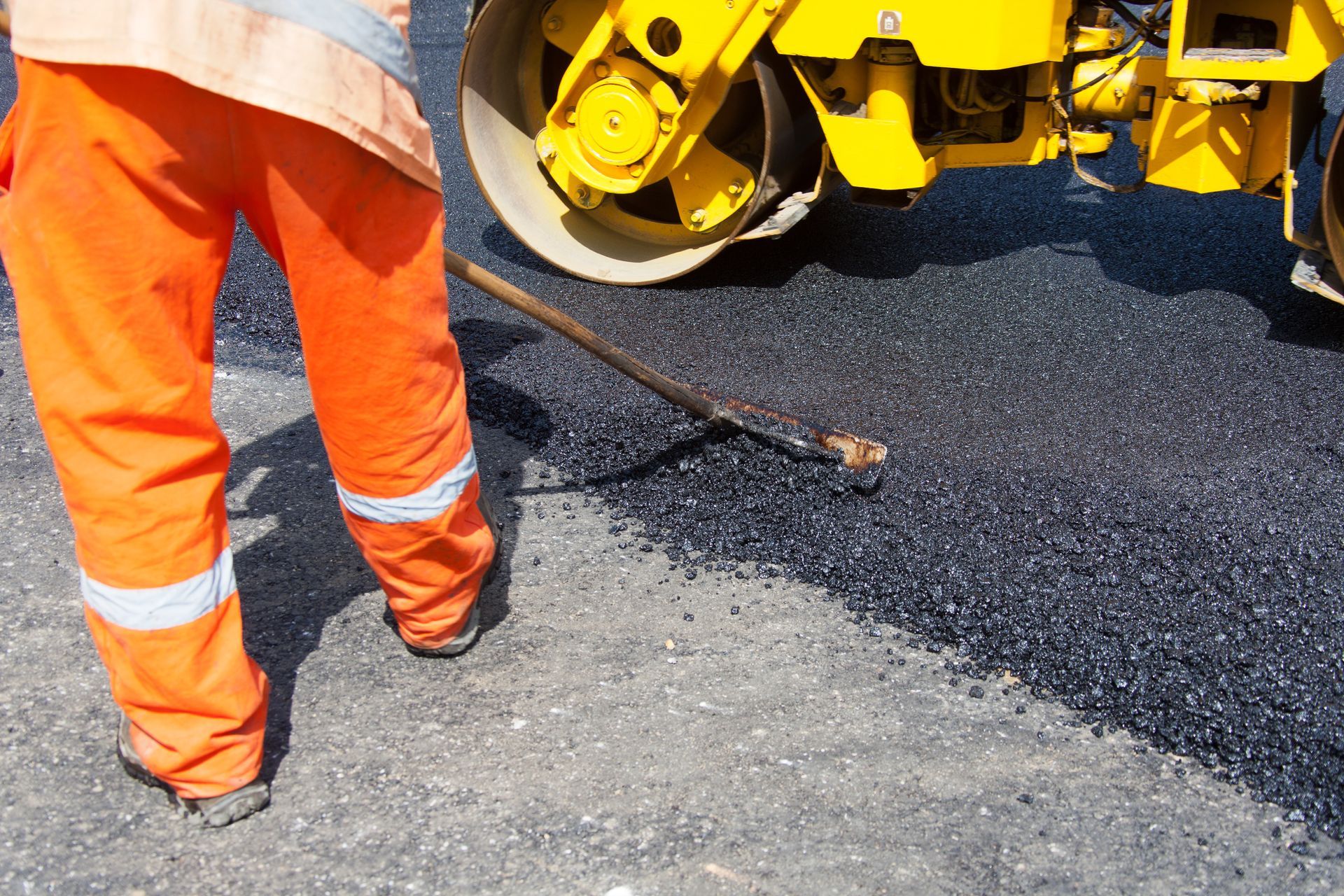 Construction worker in orange pants and safety vest uses a tool on fresh asphalt, next to a yellow road roller. Construction worker in orange pants and safety vest uses a tool on fresh asphalt, next to a yellow road roller.