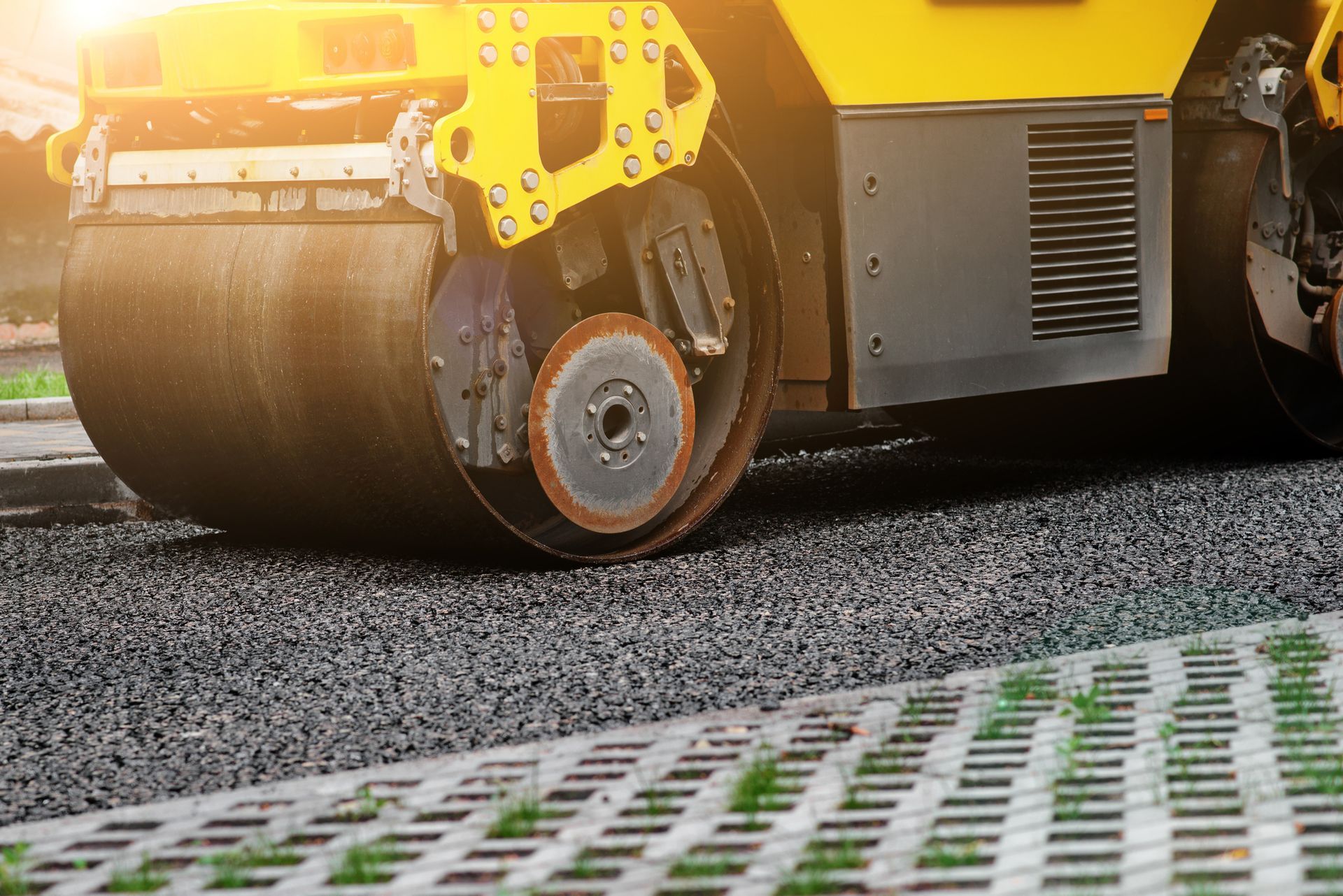 Road roller compacting fresh asphalt on a road; yellow machine, gray pavement, and green grass visible.