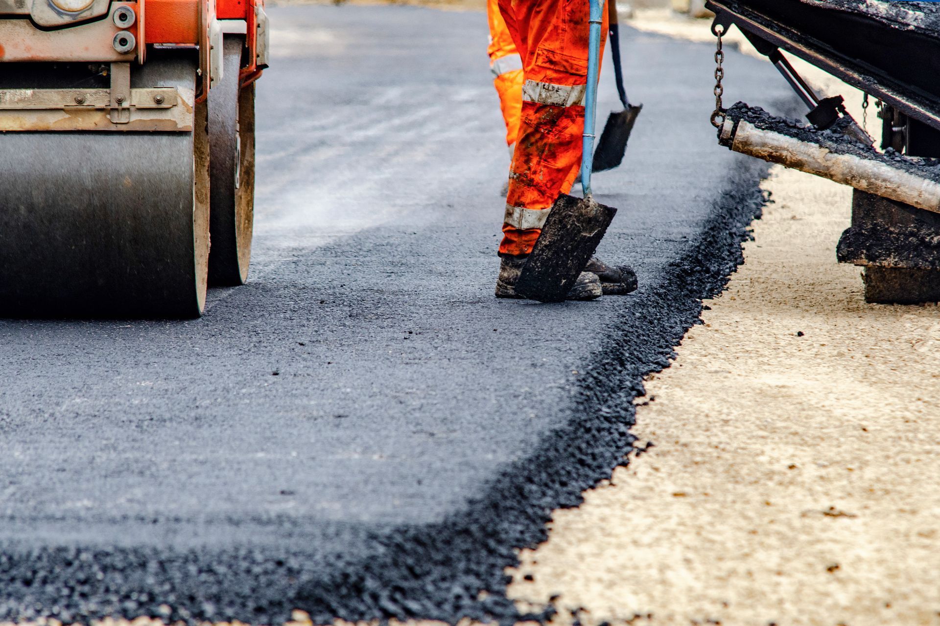 Asphalt paving contractors in high-visibility gear are applying fresh hot asphalt to a road surface.