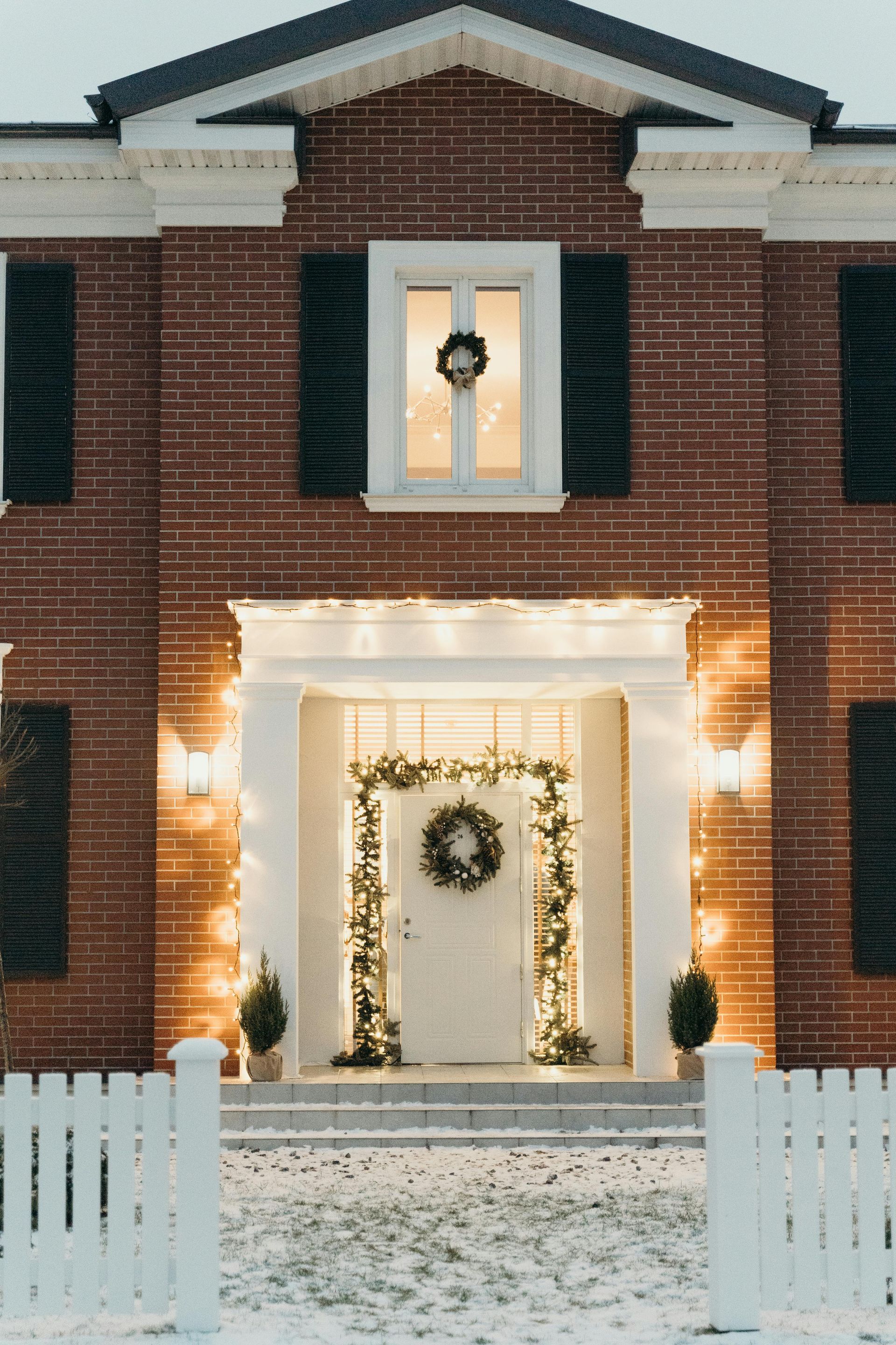 Brick house with Christmas decorations, front door framed by garland, illuminated, snow on the ground.