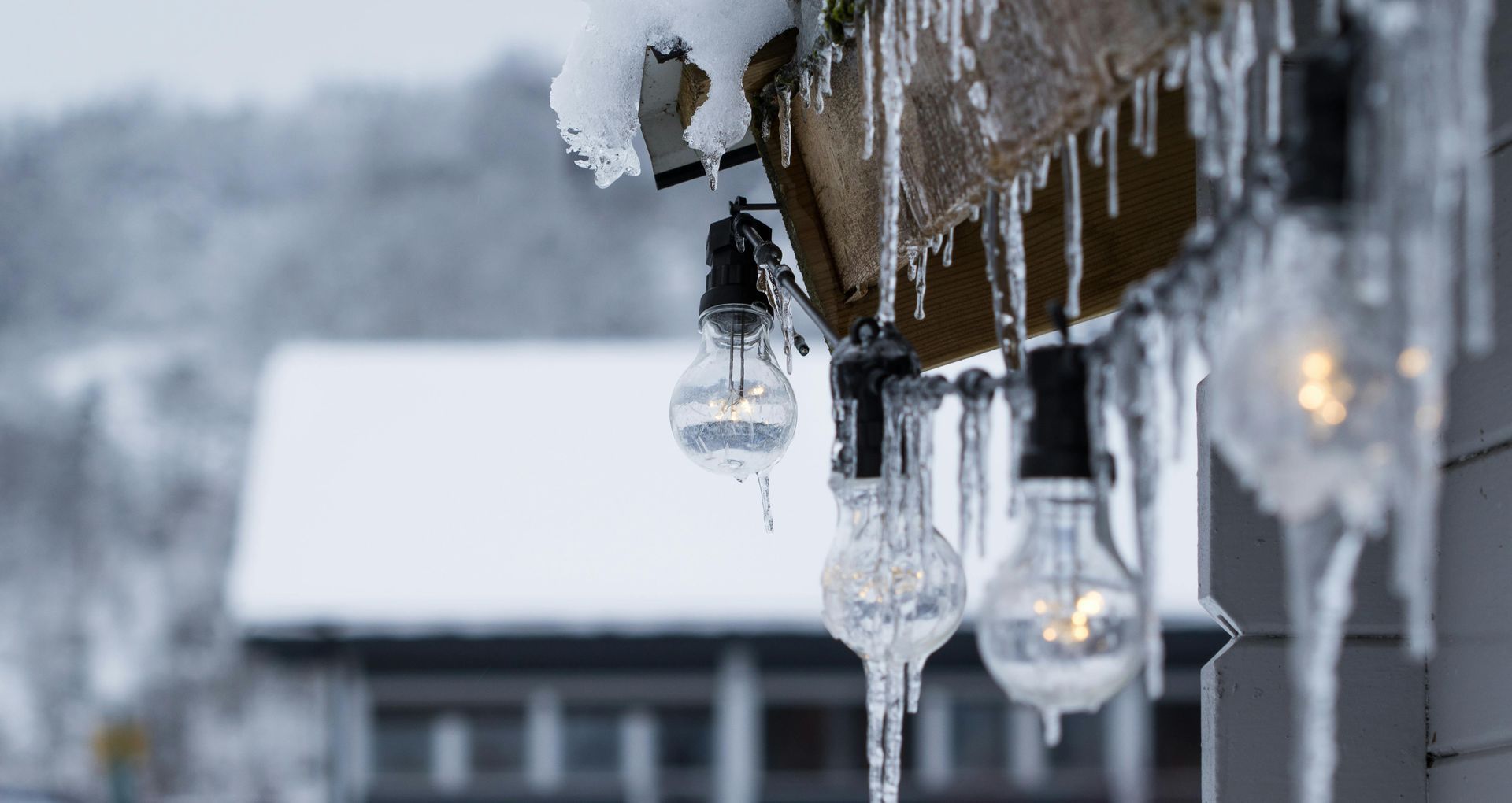 Icicles hanging from a wooden beam with lit string lights; snow-covered building in background.