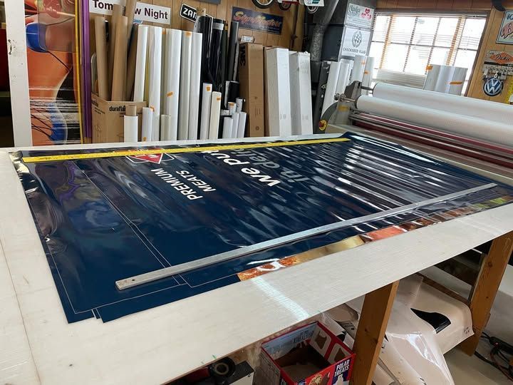 Large blue and gold sign on a table, being worked on. A ruler, window, and supplies are in the background.