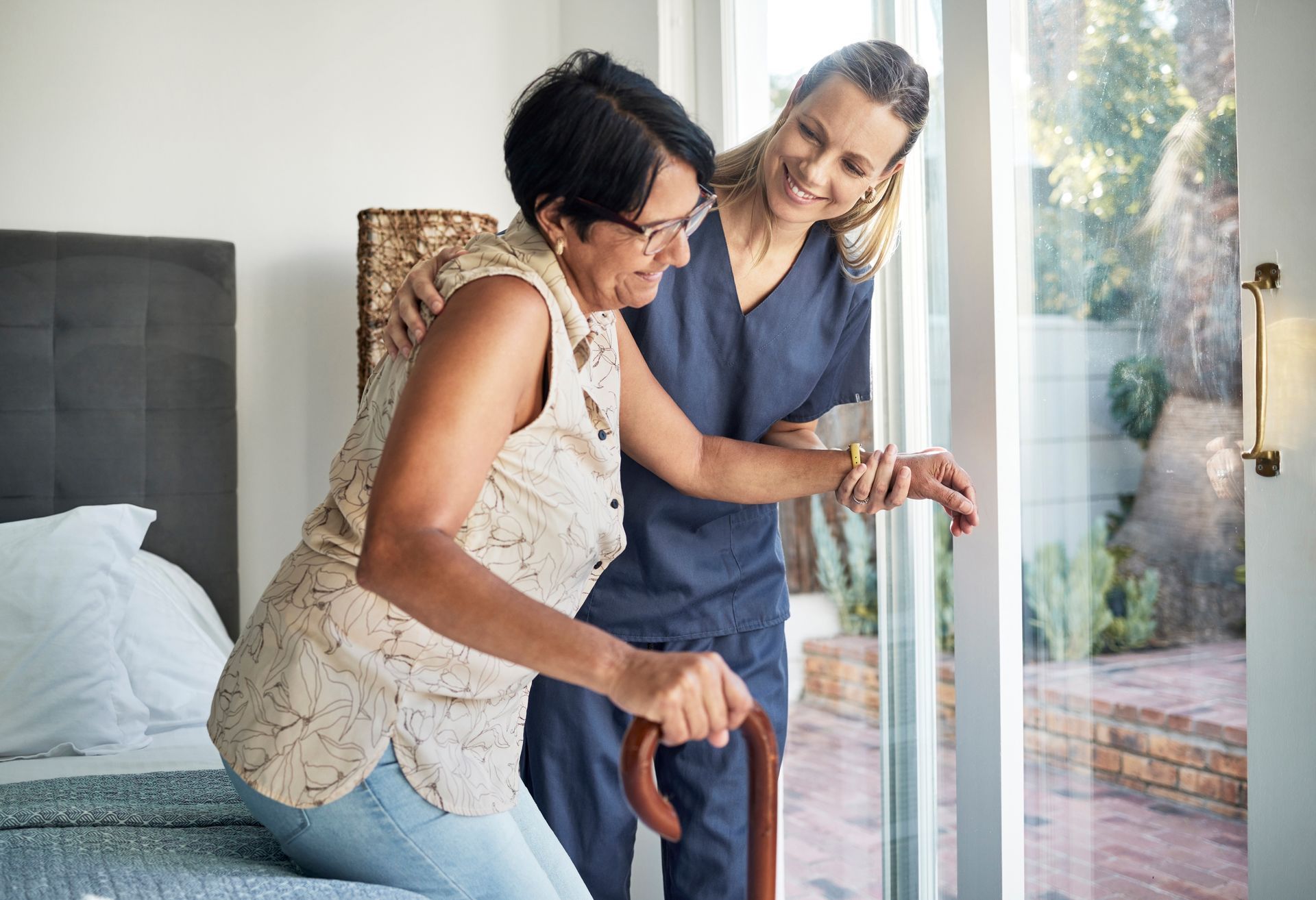 Two women help steady a cane beside a bed near a bright window