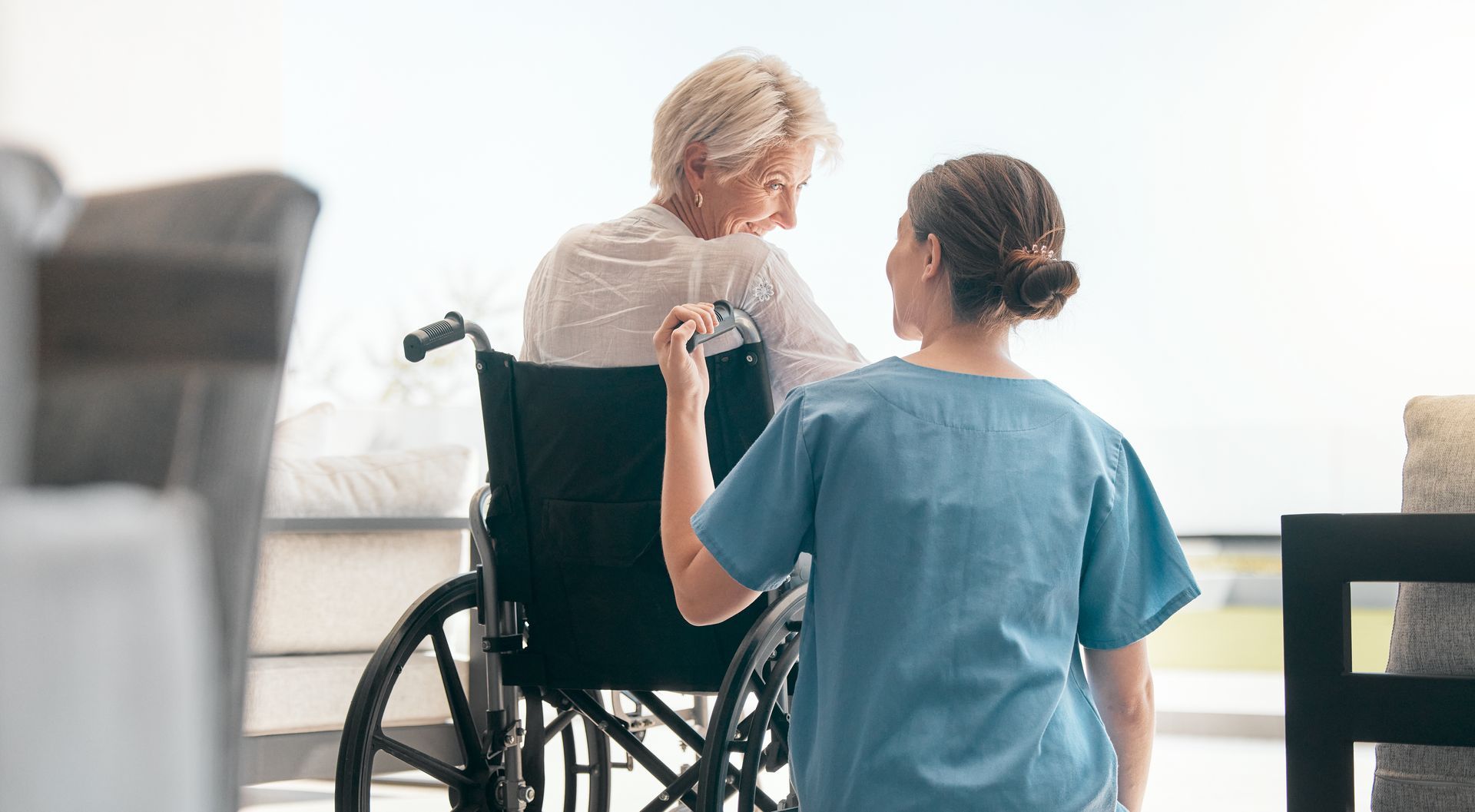 Caregiver in blue scrubs stands behind an elderly person seated in a wheelchair in a bright room