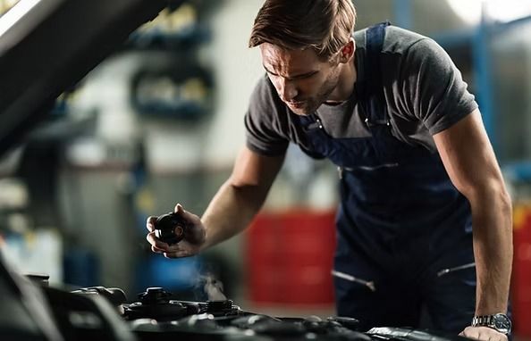 Mechanic in blue overalls examines a car engine in a shop. He looks focused and is holding a black cap. | Scottsdale Protech