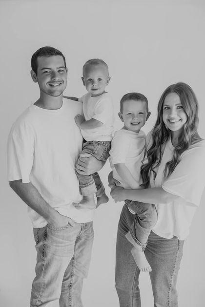 Family of four smiling, posing for a portrait; two children held by parents, wearing white shirts and jeans.