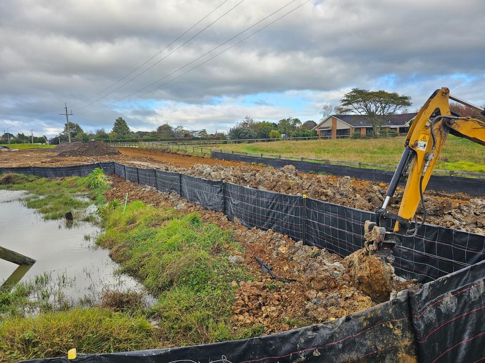 Excavator digging in an earthen containment area with standing water, under a cloudy sky.