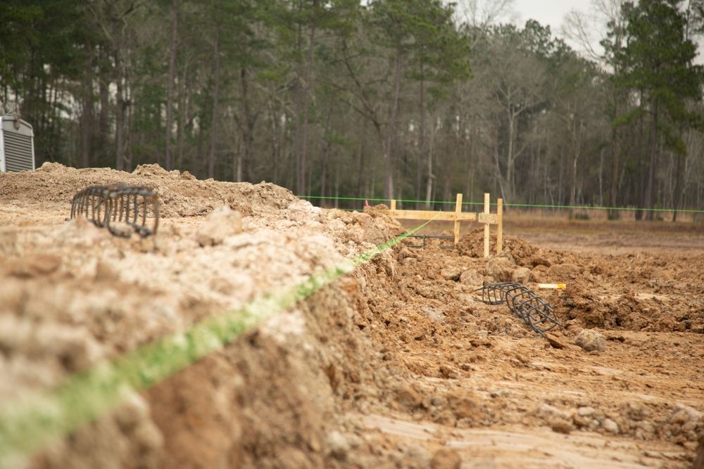 Construction site with exposed dirt, rebar, and green string line, trees in background.
