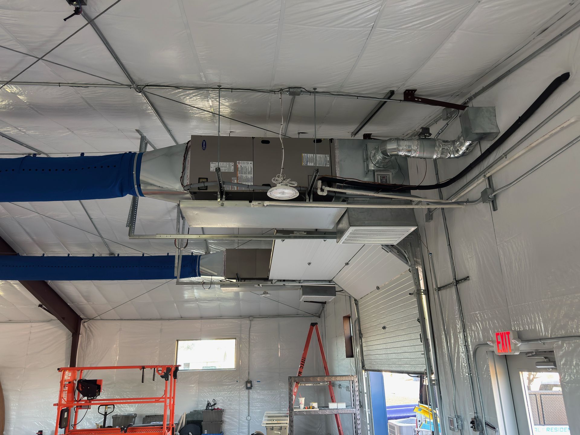 Industrial HVAC unit suspended from a white ceiling with blue fabric air ducts in a large, unfinished building.