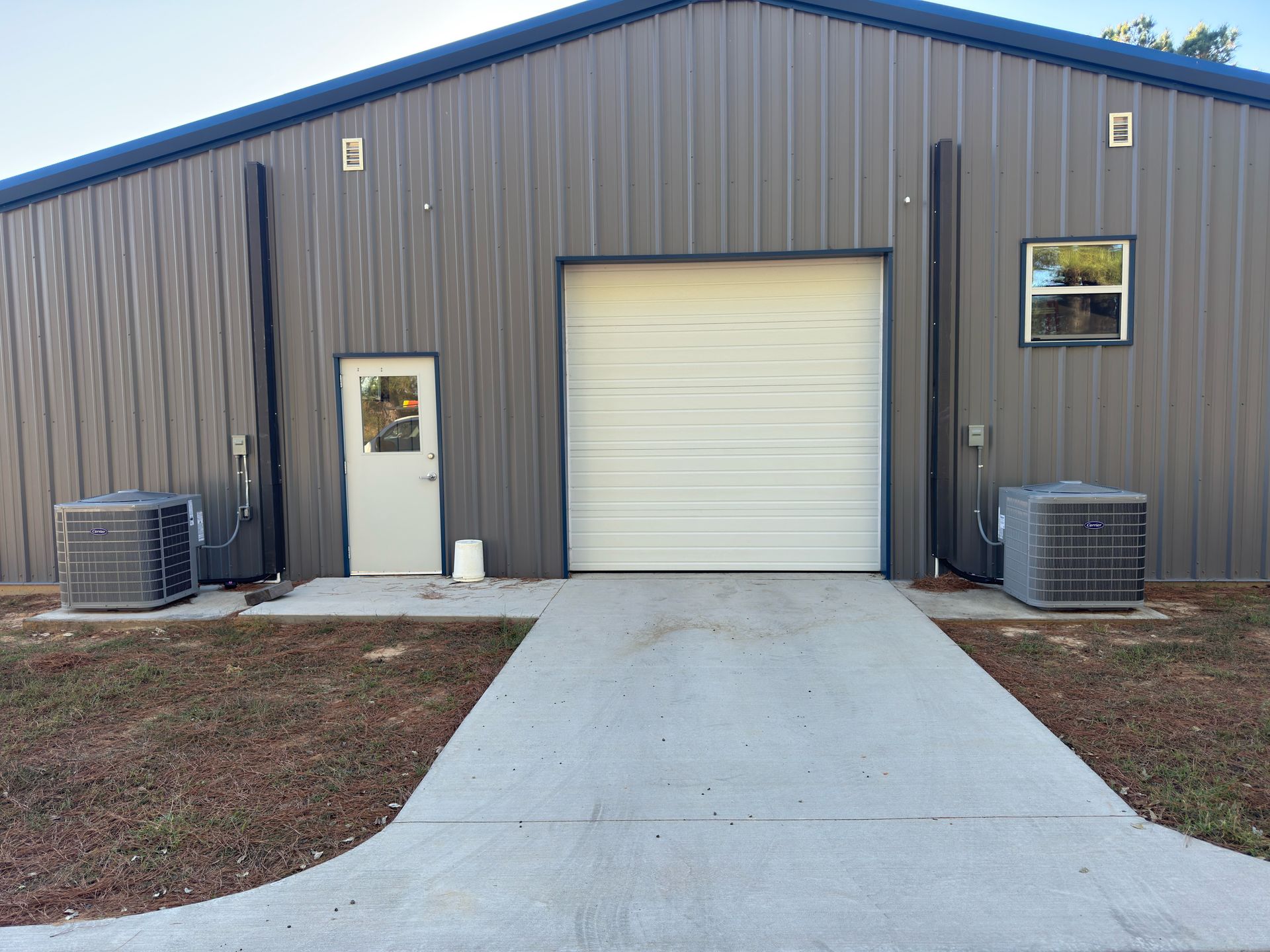 A gray metal warehouse building with a white rollup door, a single pedestrian door, and two outdoor AC units.
