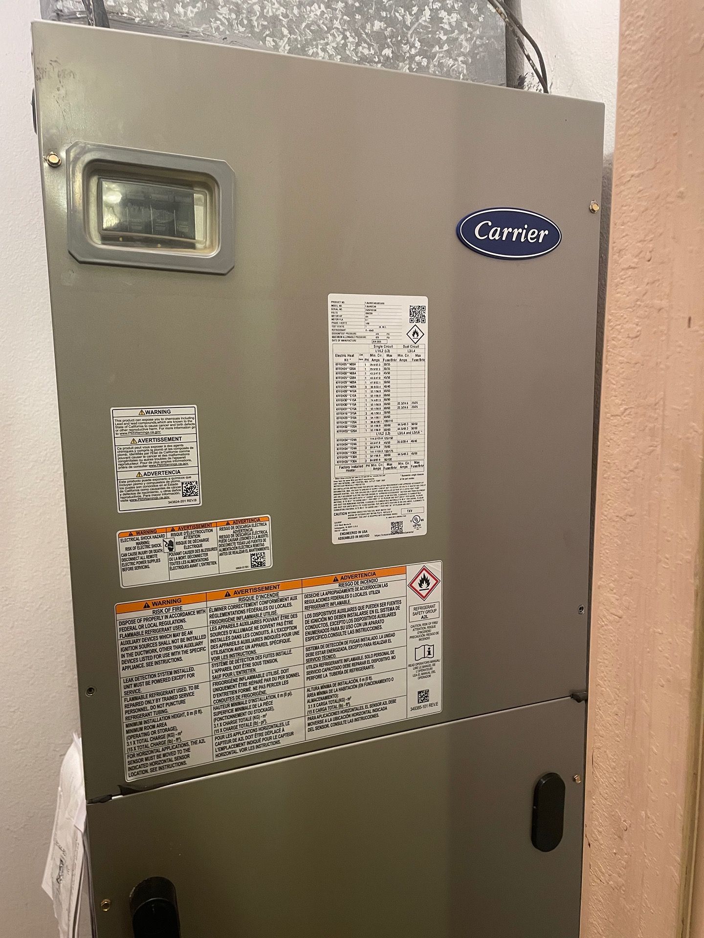 A gray Carrier HVAC unit mounted indoors against a white wall with several informational stickers on its front panel.