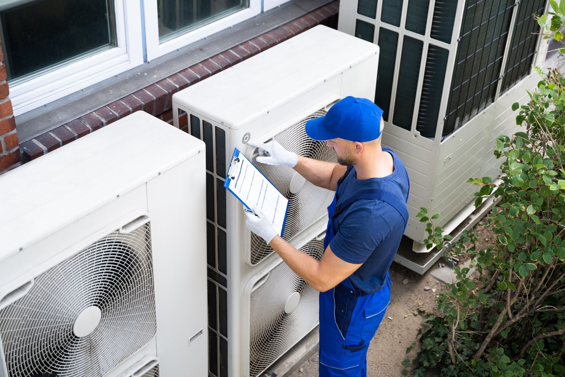 HVAC technician inspecting air conditioning units outside a building, wearing blue uniform and holding clipboard. HVAC technician inspecting air conditioning units outside a building, wearing blue uniform and holding clipboard.