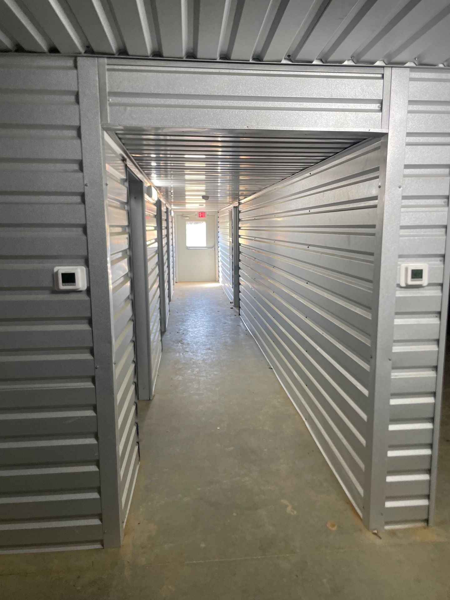 A dimly lit, narrow corridor in a storage facility with gray corrugated metal walls and a light at the far end.