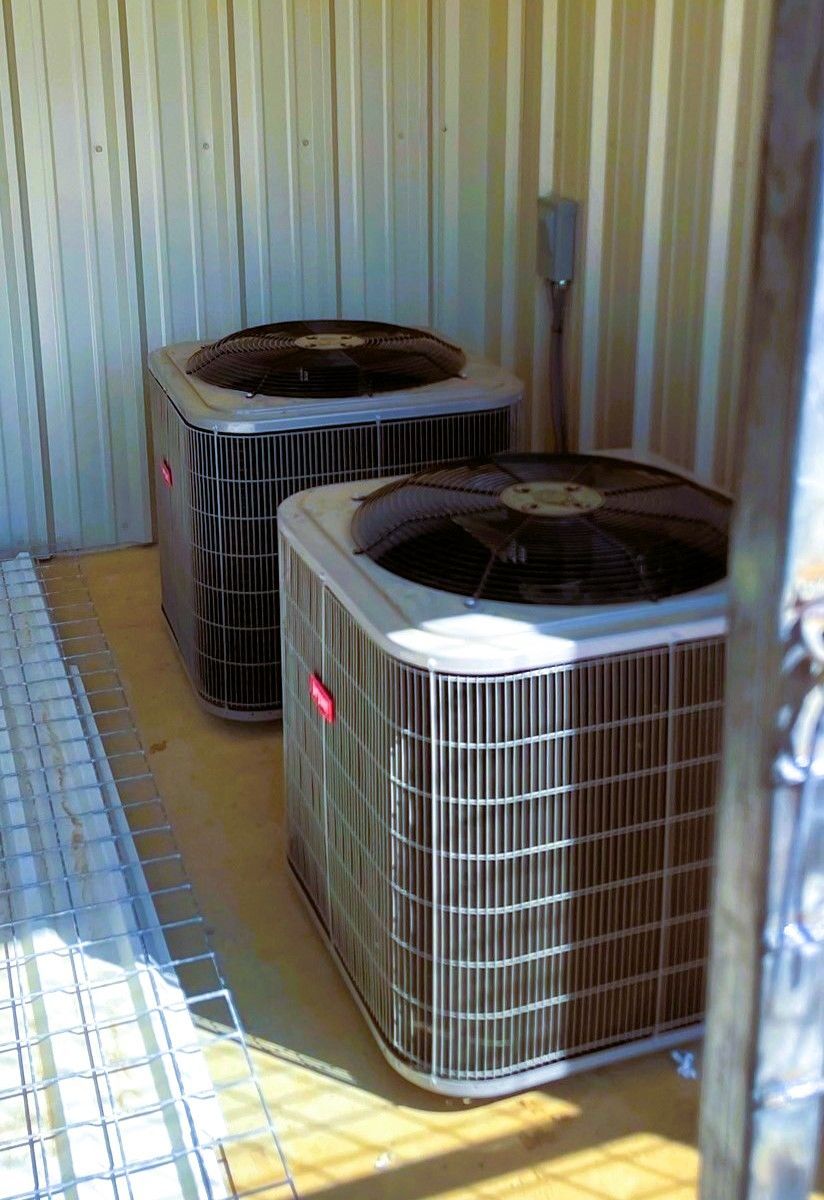 Two rectangular HVAC condenser units placed side-by-side in a narrow alcove with corrugated metal walls.