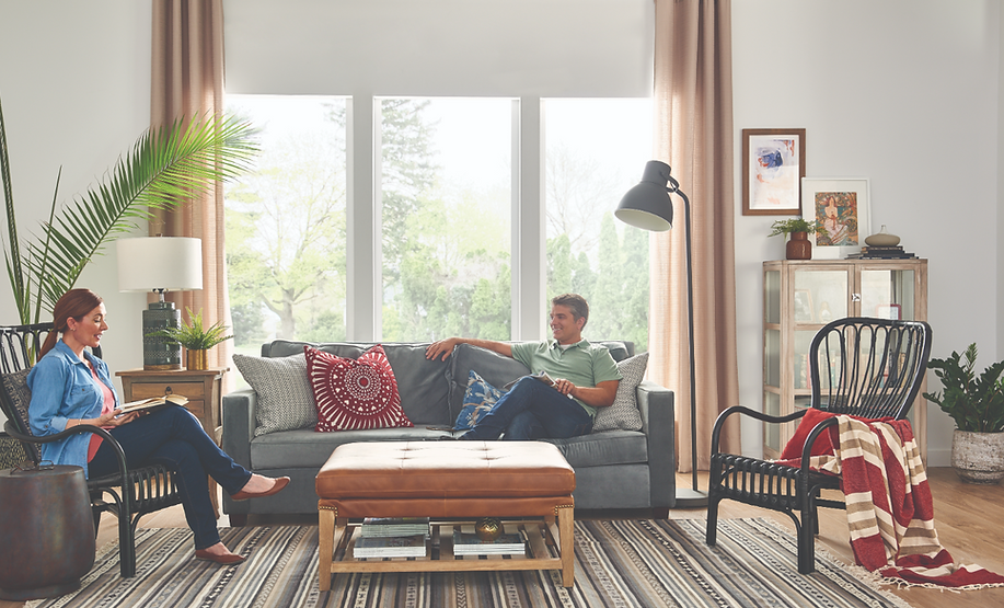 Couple in a living room, seated and relaxing. Gray sofa, large window, potted plants, floor lamp.