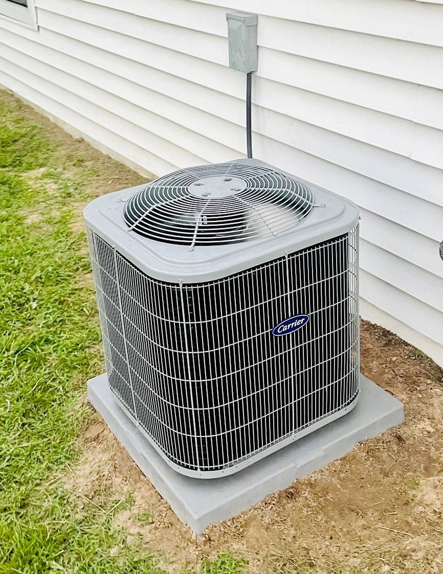 A grey outdoor air conditioning unit sits on a concrete pad against white vinyl siding next to a grassy lawn.