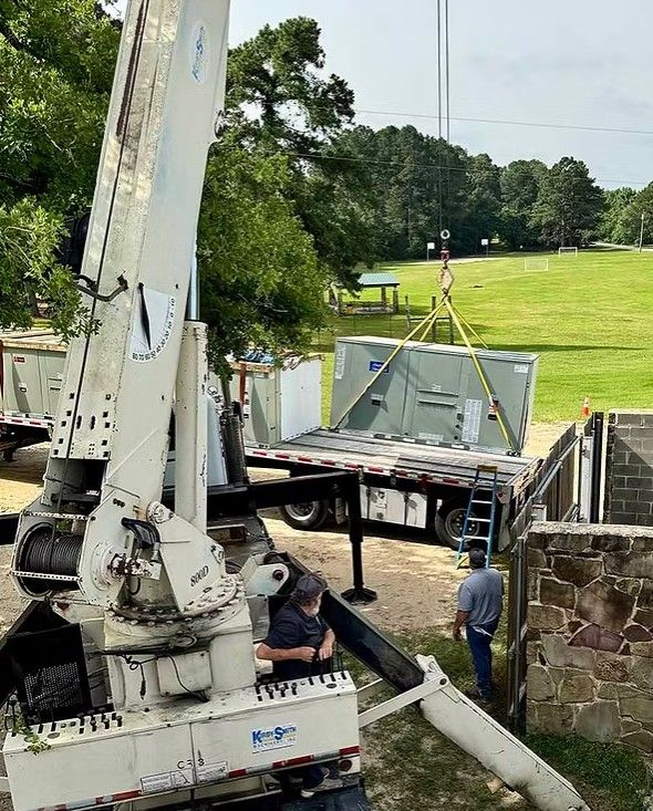 A crane lifts a large industrial HVAC unit from a flatbed truck near a stone wall in a grassy field.
