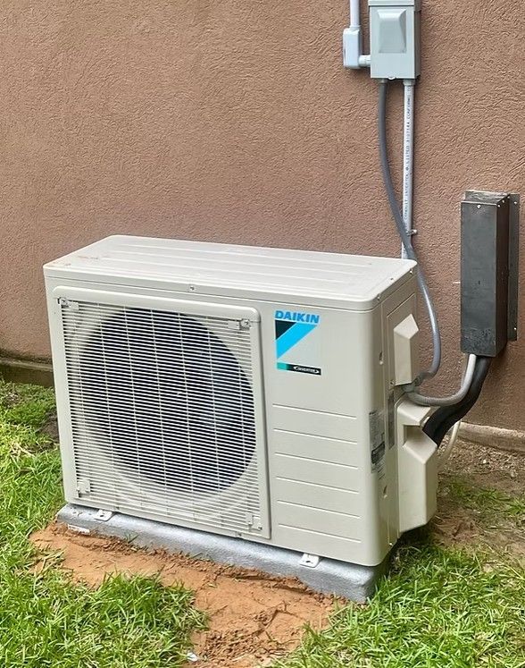 A Daikin outdoor HVAC unit mounted on a concrete pad against a brown wall with electrical wiring.