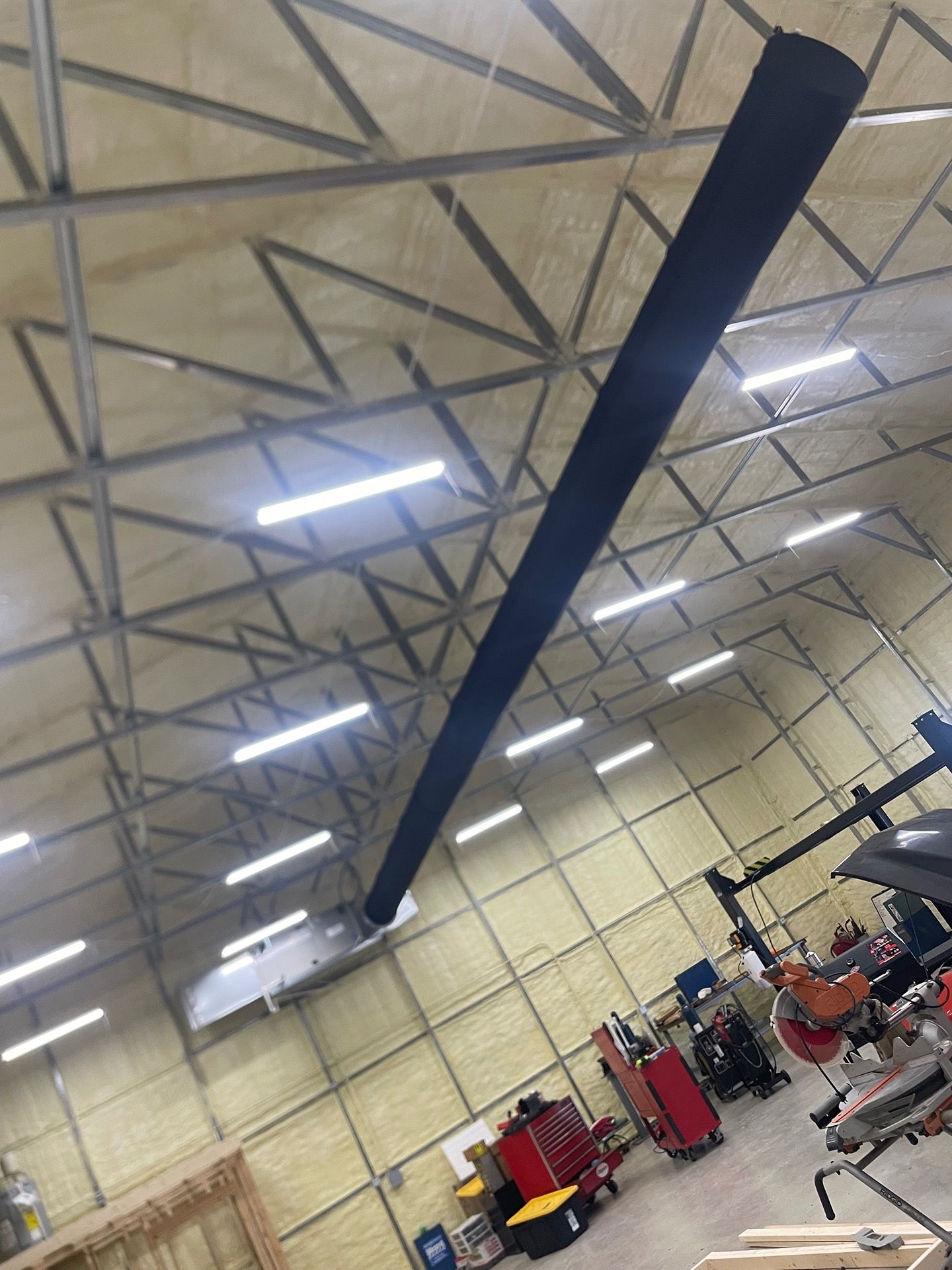 A long, black fabric air duct hangs from the ceiling rafters in a large, workshop-style building with LED lighting.