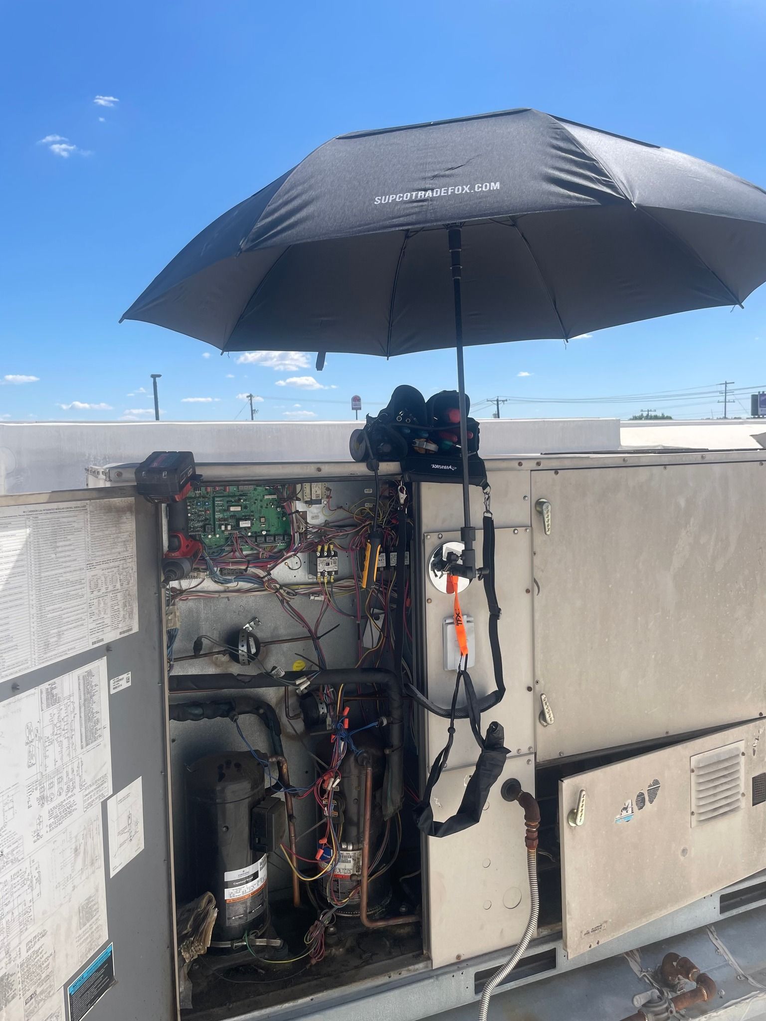 A black umbrella is strapped to an open rooftop HVAC unit to provide shade while it is being serviced.