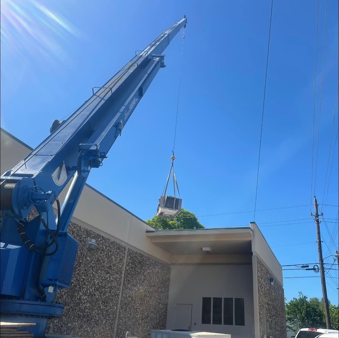 A blue crane lifting an HVAC unit onto the roof of a stone-walled commercial building under a clear blue sky.