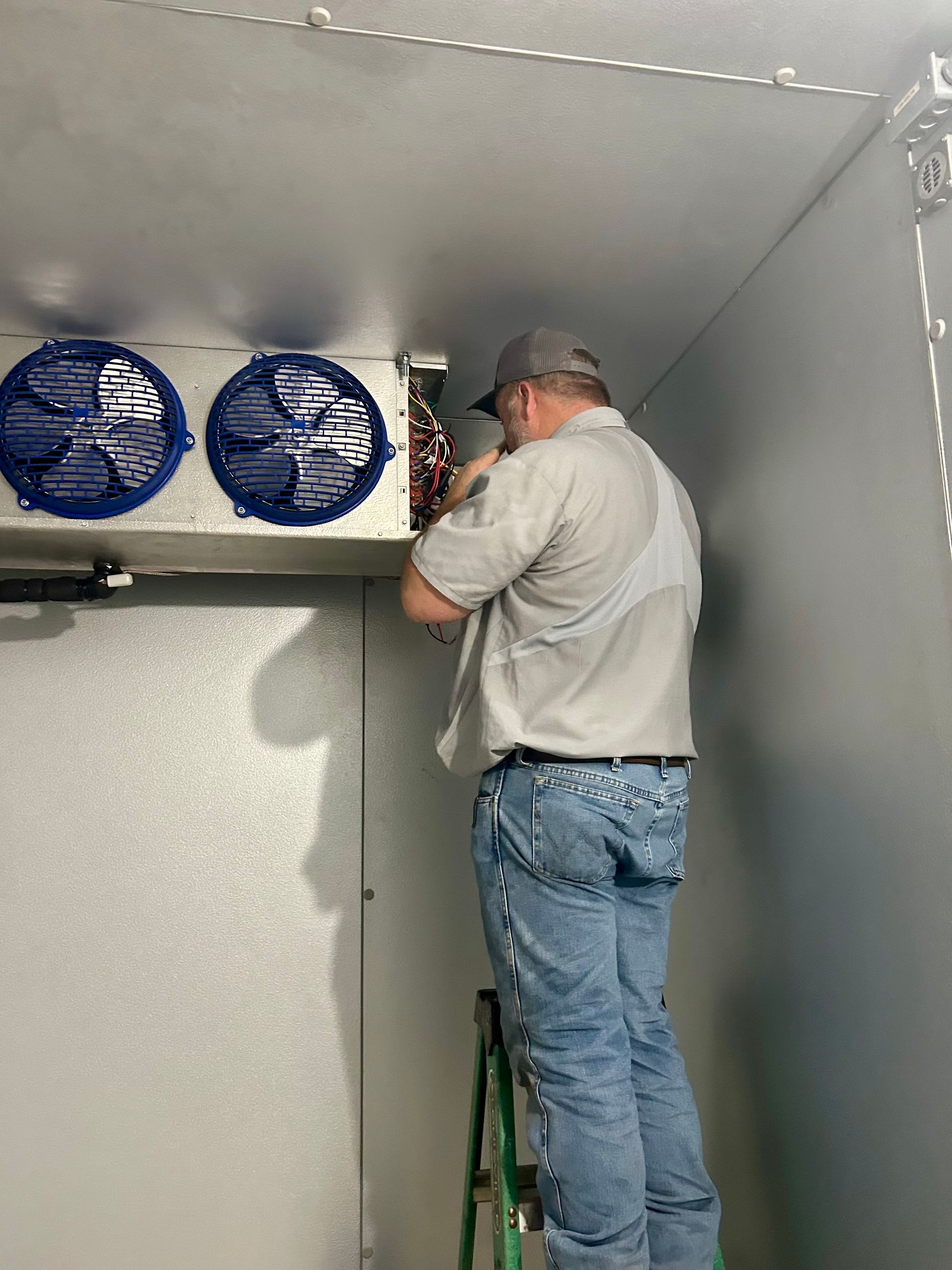 A technician on a ladder repairs an industrial refrigeration unit mounted to the ceiling of a white-walled room.