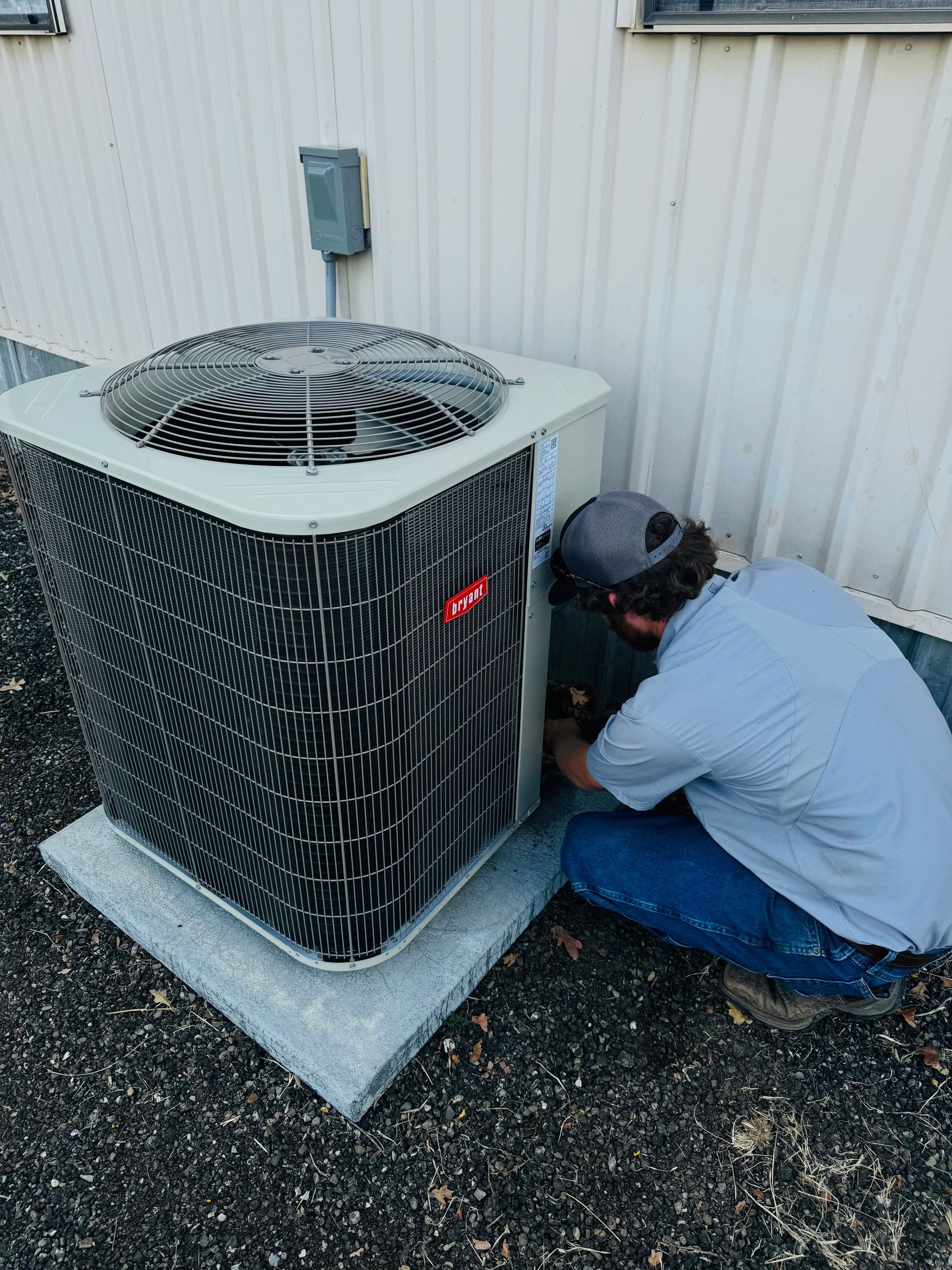 A person in a light blue shirt and baseball cap kneels while servicing an outdoor residential air conditioning unit.