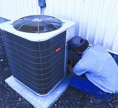 HVAC technician in red shirt, kneeling, attaching gauges to an AC unit outside a brick building.