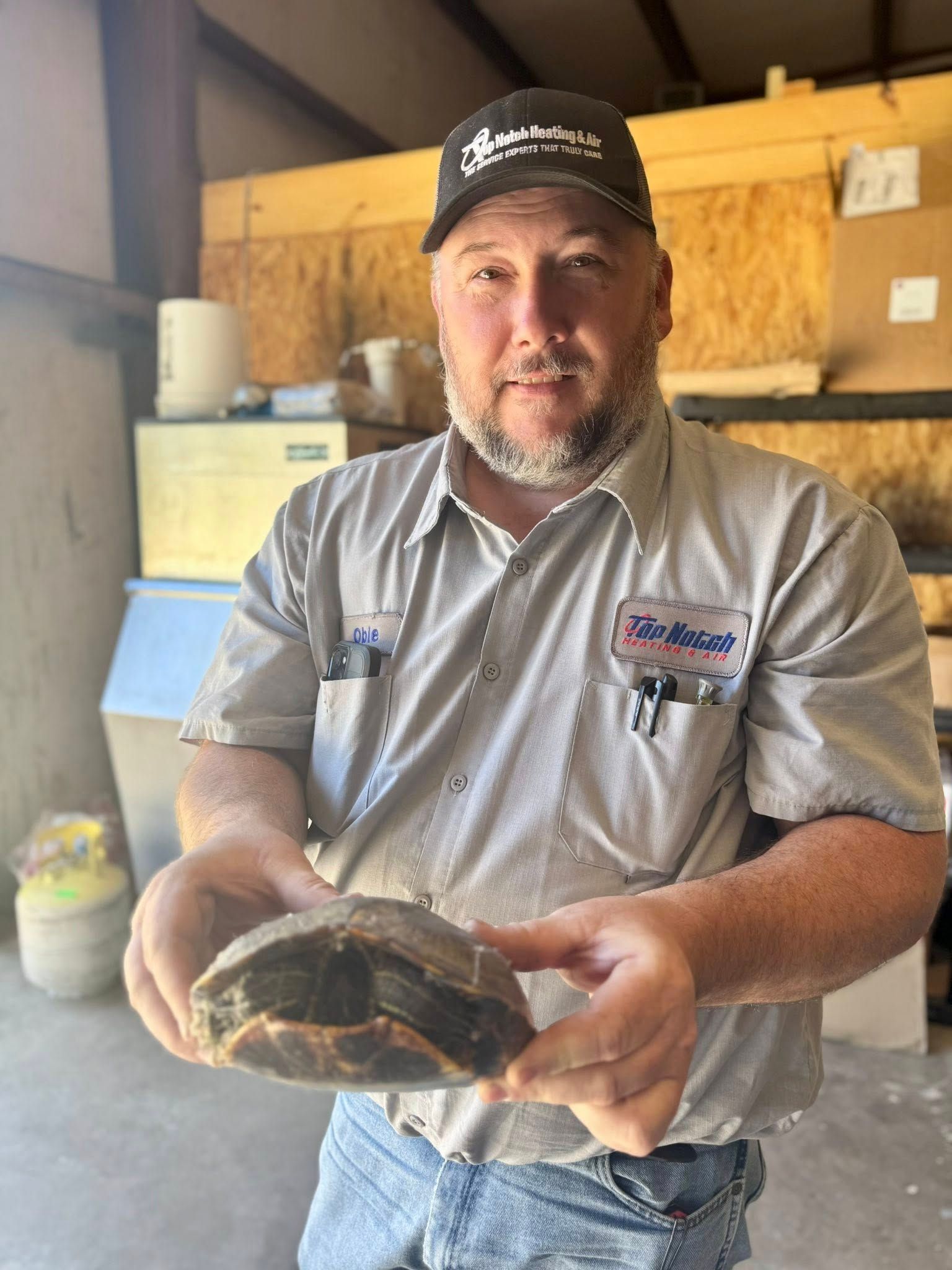 A person in a gray work shirt and cap holding a small turtle in an indoor setting.