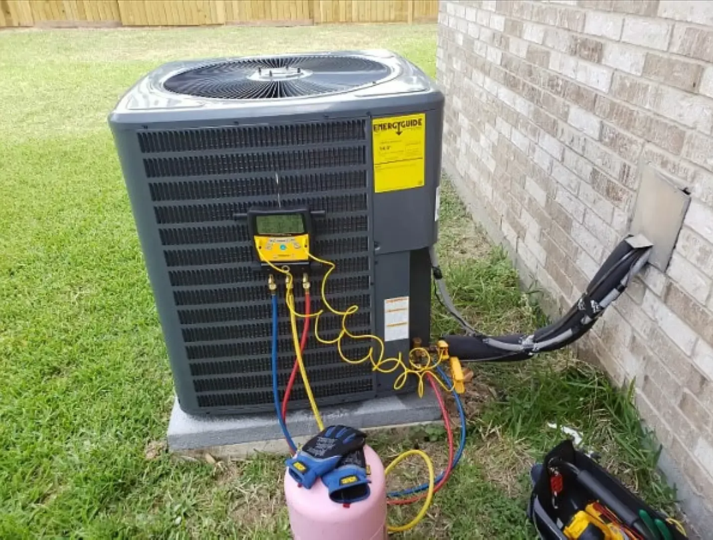 A technician is using a digital gauge manifold to service an outdoor residential air conditioning unit on a grassy lawn.