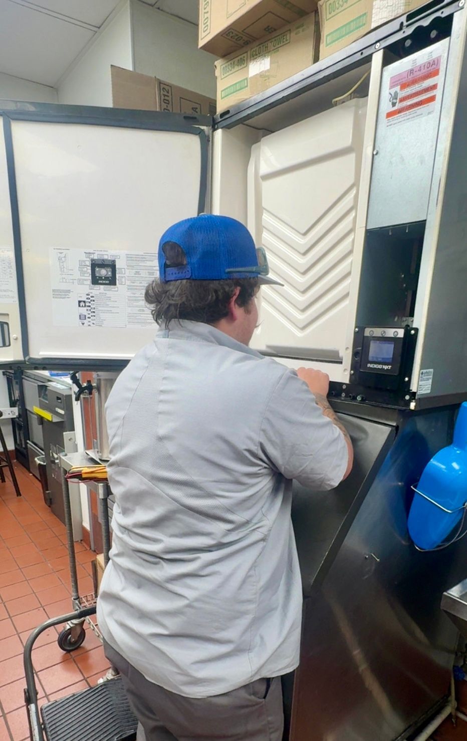 A person wearing a blue cap and grey shirt works on a large, open ice machine in a commercial kitchen. A person wearing a blue cap and grey shirt works on a large, open ice machine in a commercial kitchen.