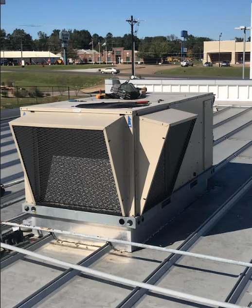 A tan commercial HVAC rooftop unit sits on a metal roof under a clear sky, with tools resting on its top.