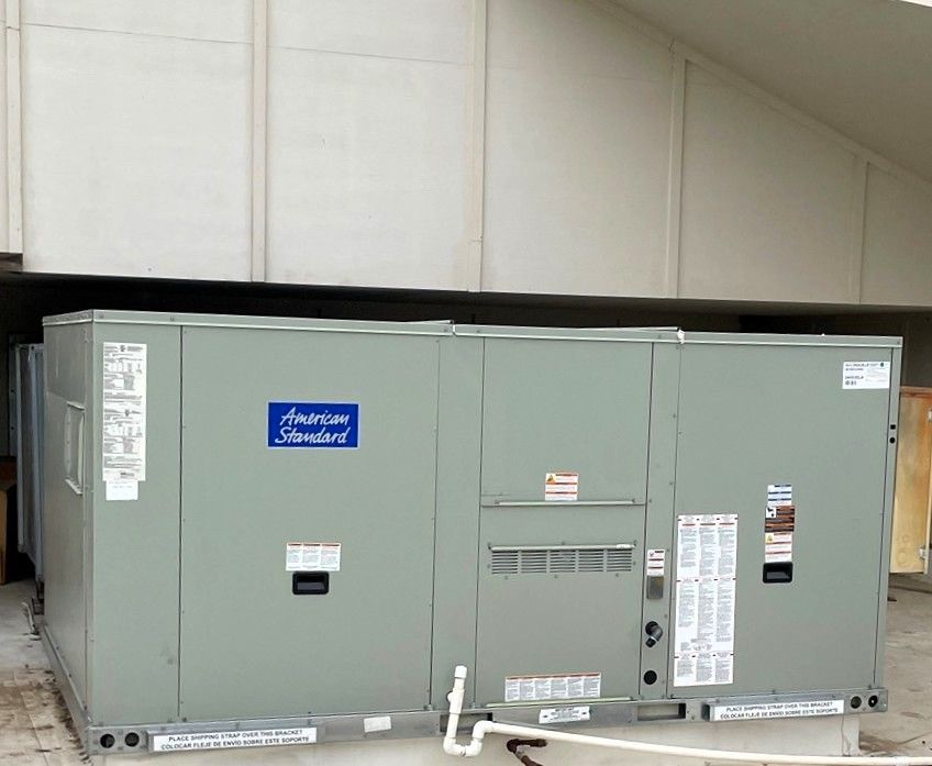 A light green American Standard commercial rooftop HVAC unit sits on a roof under an overhang.